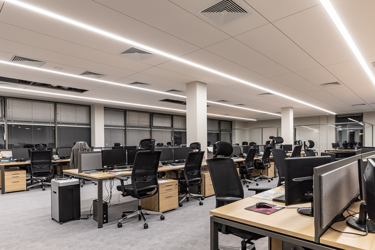 Nowoczesne biuro z rzędami biurek, czarnymi krzesłami obrotowymi i oświetleniem LED w długich pasach na suficie. Widoczne komputery, segregatory i rolety w oknach.