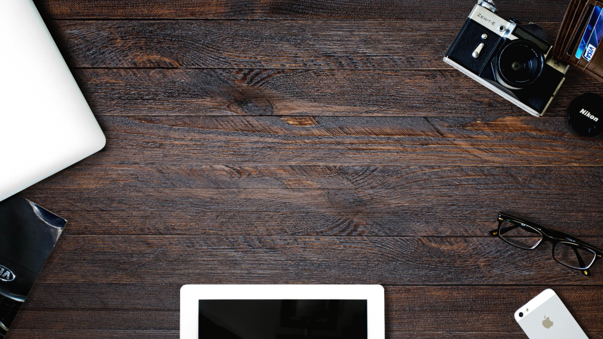 Dark wooden desk surface with a laptop, tablet, vintage Zenit-E camera, glasses, smartphone, wallet with a Visa card, and a Nikon lens cap, arranged for a clean, organized workspace aesthetic.