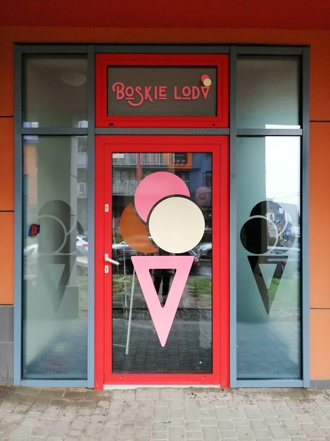 Oklejona witryna lodziarni z grafiką lodów w wafelku, wykonana z folii plotowanej, z nazwą 'Boskie Lody' nad drzwiami.