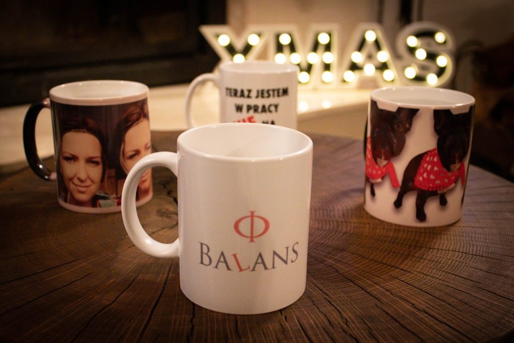 Cztery białe kubki z różnymi nadrukami stojące na drewnianym blacie: jeden z portretem dwóch kobiet, drugi z napisem 'Teraz jestem w pracy', trzeci z wizerunkiem psów w sweterkach, a czwarty z logo...