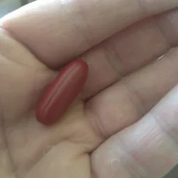 Podłużna, czerwona kapsułka leży na otwartej dłoni, widoczne linie papilarne.