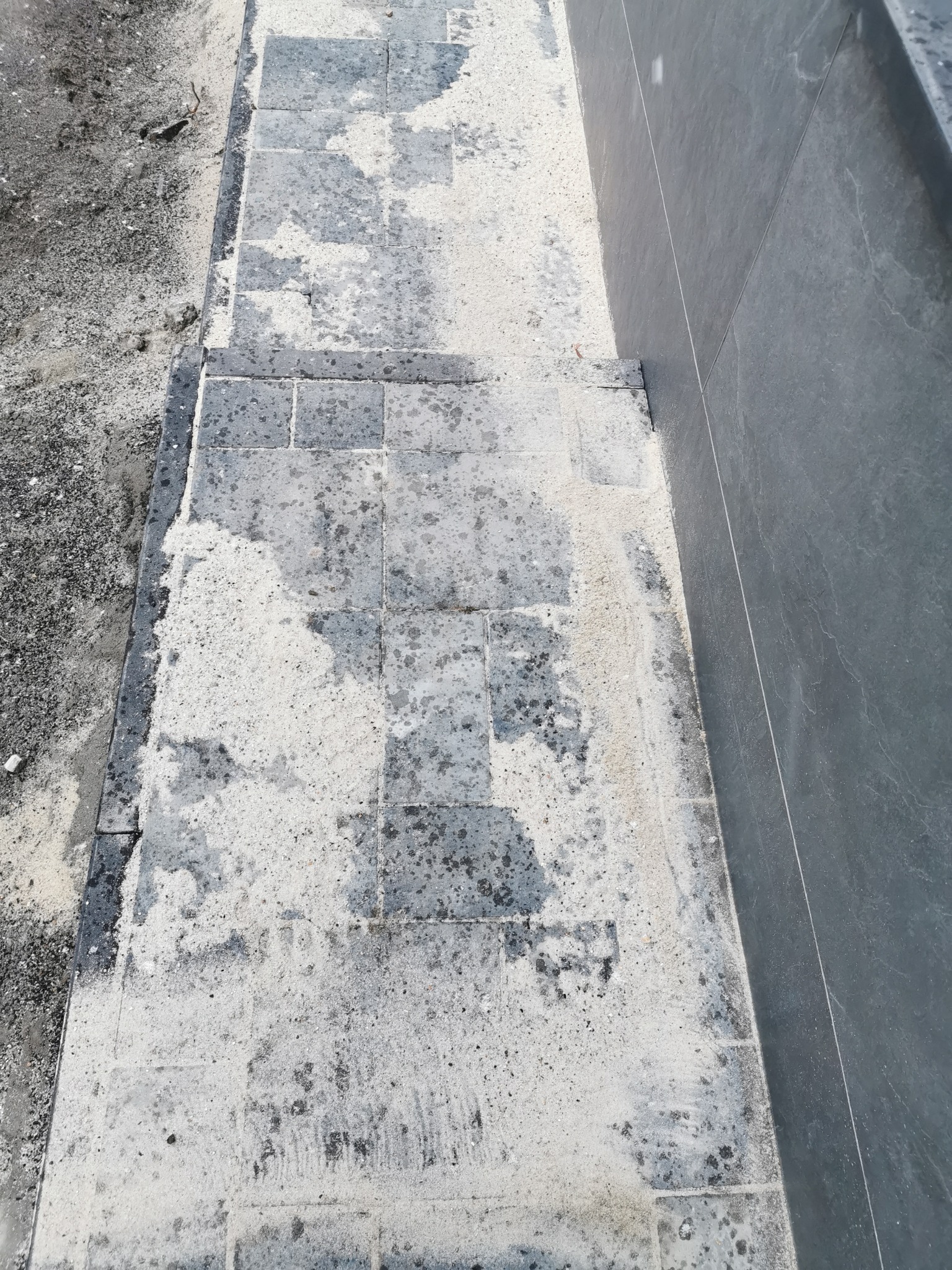 Świeżo ułożona, szara kostka brukowa z pozostałościami piasku kwarcowego do fugowania, przylegająca do ciemnej ściany budynku.