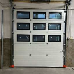 TESSA-DOM KAROL TESSA SPÓŁKA KOMANDYTOWA - Biała segmentowa brama garażowa z prostokątnymi okienkami, widoczne elementy montażowe i sprężyna nad bramą.