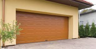 Zamknięta, brązowa brama garażowa segmentowa z poziomymi przetłoczeniami, zamontowana w elewacji budynku o jasnym, piaskowym kolorze. Z lewej strony widoczny mały krzew, a w tle iglaki i fragment...