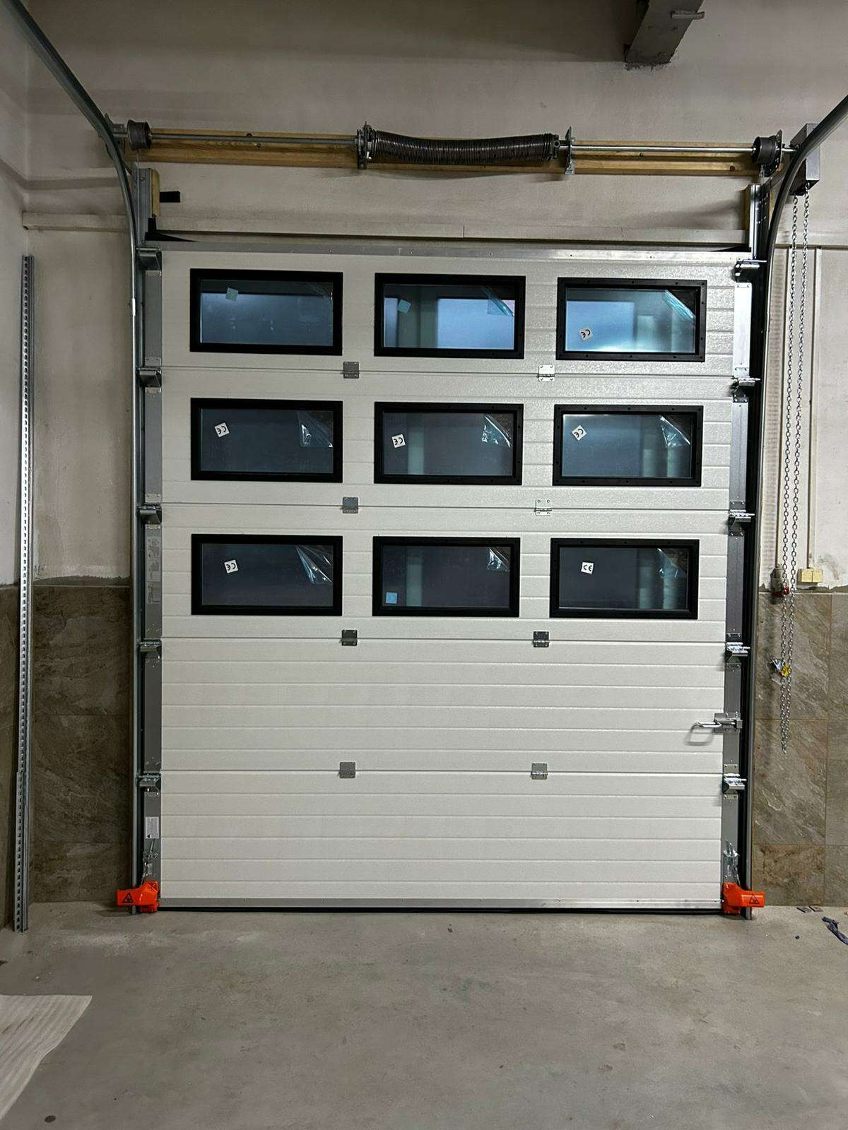 Biała segmentowa brama garażowa z prostokątnymi okienkami, widoczne elementy montażowe i sprężyna nad bramą.