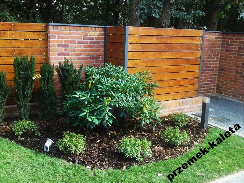 Nowoczesne ogrodzenie drewniane z elementami murowanymi, wkomponowane w aranżację ogrodu z roślinami i oświetleniem punktowym.