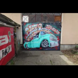 Graffiti na metalowej bramie garażowej przedstawiające turkusowy samochód typu pickup z napisem 'Auto Mechanik' i numerem telefonu. Widoczne otoczenie garażu i fragment reklamy.