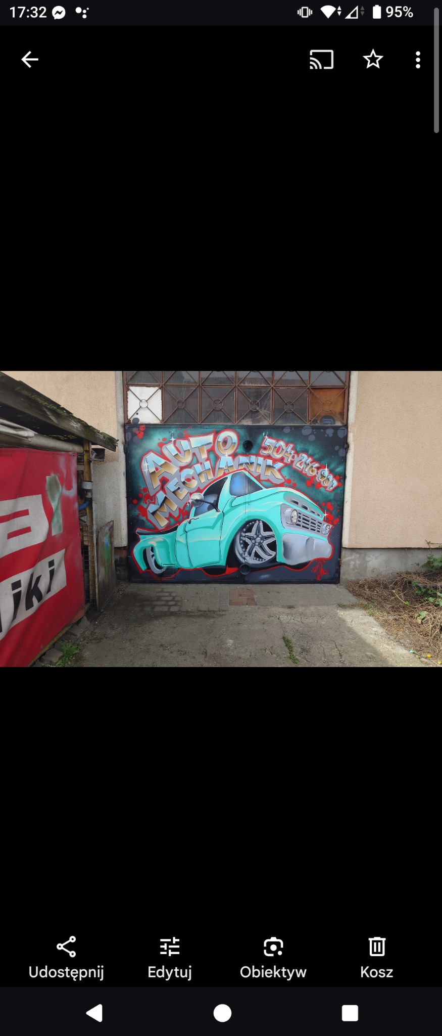 Graffiti na metalowej bramie garażowej przedstawiające turkusowy samochód typu pickup z napisem 'Auto Mechanik' i numerem telefonu. Widoczne otoczenie garażu i fragment reklamy.