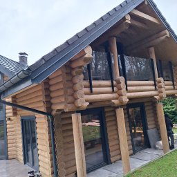 Wikpol sp. z o.o. - Dwukondygnacyjny dom z bali z ciemną dachówką, balkonem z czarnymi balustradami i szarymi oknami, widok z zewnątrz na szarym bruku i zielonym trawniku.