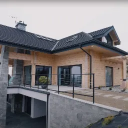 Nowoczesny dom z bali z elementami kamiennymi, tarasem i pergolą na zboczu wzgórza, z widokiem na zalesioną dolinę w pochmurny dzień.