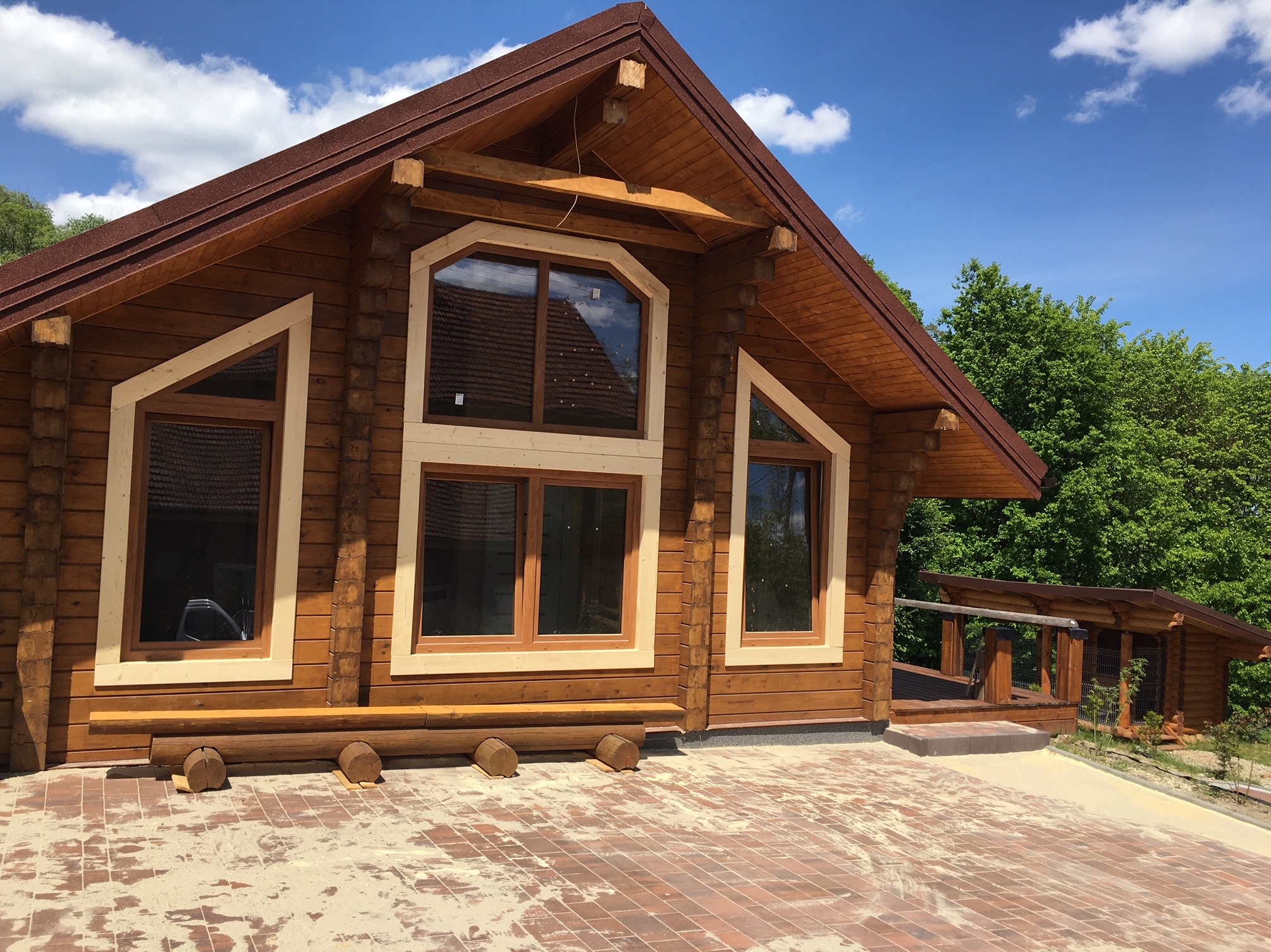 Fasada domu z bali klejonej z charakterystycznymi oknami o nietypowych kształtach, widoczna drewniana ławka przed domem i fragment brukowanej powierzchni, błękitne niebo w tle.