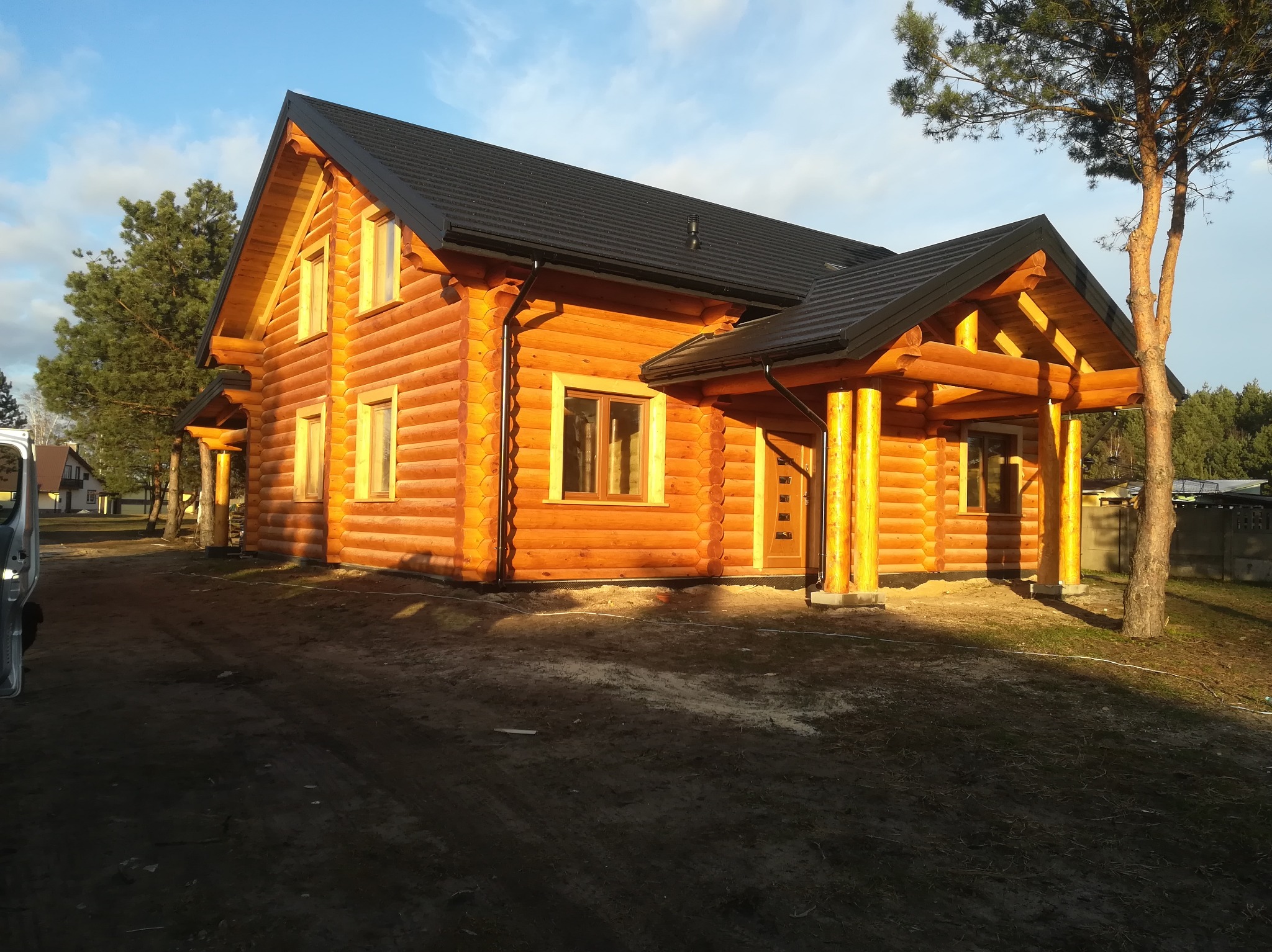 Nowoczesny dom z bali, z czarnym dachem i drewnianą werandą, otoczony drzewami, z widocznym fragmentem samochodu po lewej stronie kadru.