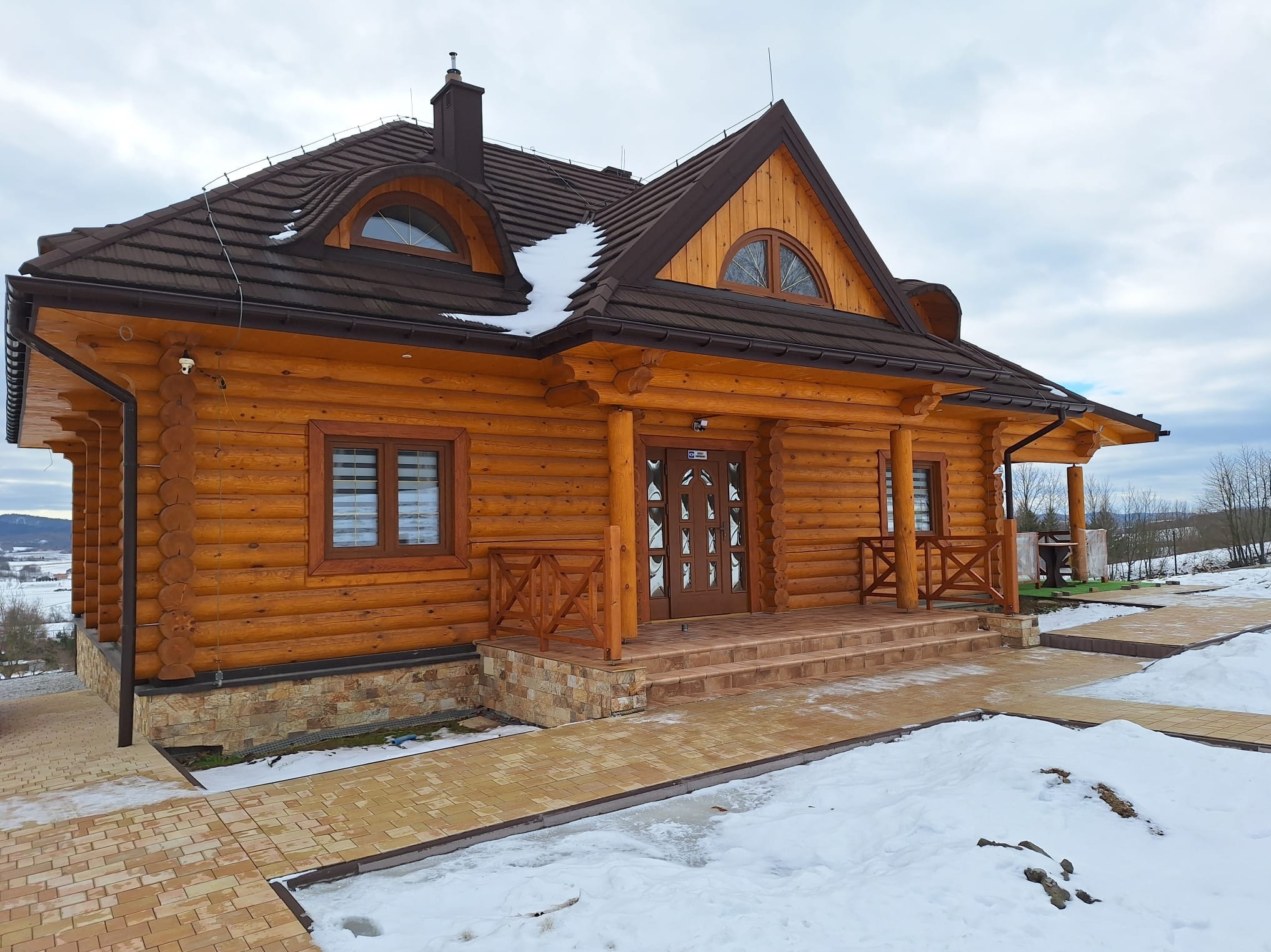 Nowoczesny dom z bali z brązowym dachem i elementami dekoracyjnymi, otoczony śniegiem i brukowaną ścieżką.