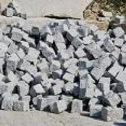 Stos szarych, obrobionych kamieni o nieregularnych kształtach, prawdopodobnie przeznaczonych do układania nawierzchni.