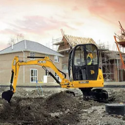 Mini koparka JCB 8030 ZTS w trakcie prac ziemnych, widoczny wykop, w tle domy jednorodzinne w trakcie budowy i lekko zachmurzone niebo o zachodzie słońca.