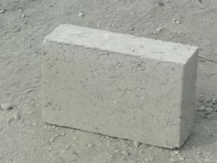 Pojedynczy, szary pustak żużlobetonowy o wymiarach 49x24x24 leżący na szarym, gruzowym podłożu.