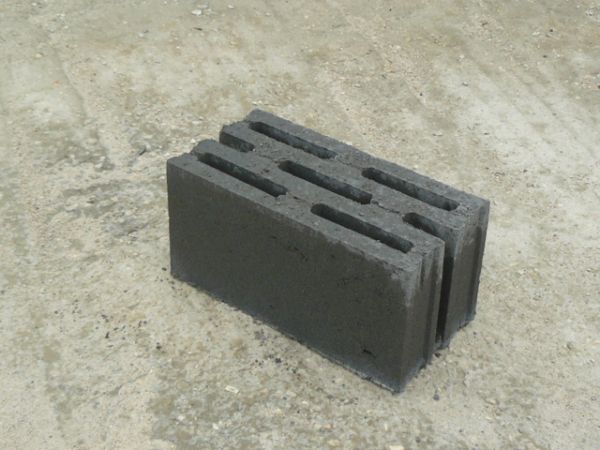 Pojedynczy blok żużlobetonowy Alfa z trzema podłużnymi otworami, leżący na betonowej powierzchni.