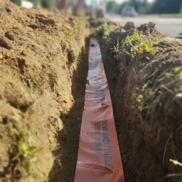 Wykop w ziemi z pomarańczową taśmą ostrzegawczą z napisem: UWAGA! KABEL OPTO-TELEKOMUNIKACYJNY. W tle rozmyty krajobraz z ulicą i koparką.