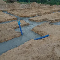 Wylane fundamenty pod budynek z widocznymi niebieskimi rurami instalacyjnymi wystającymi z betonu, otoczone piaskiem i ziemią.