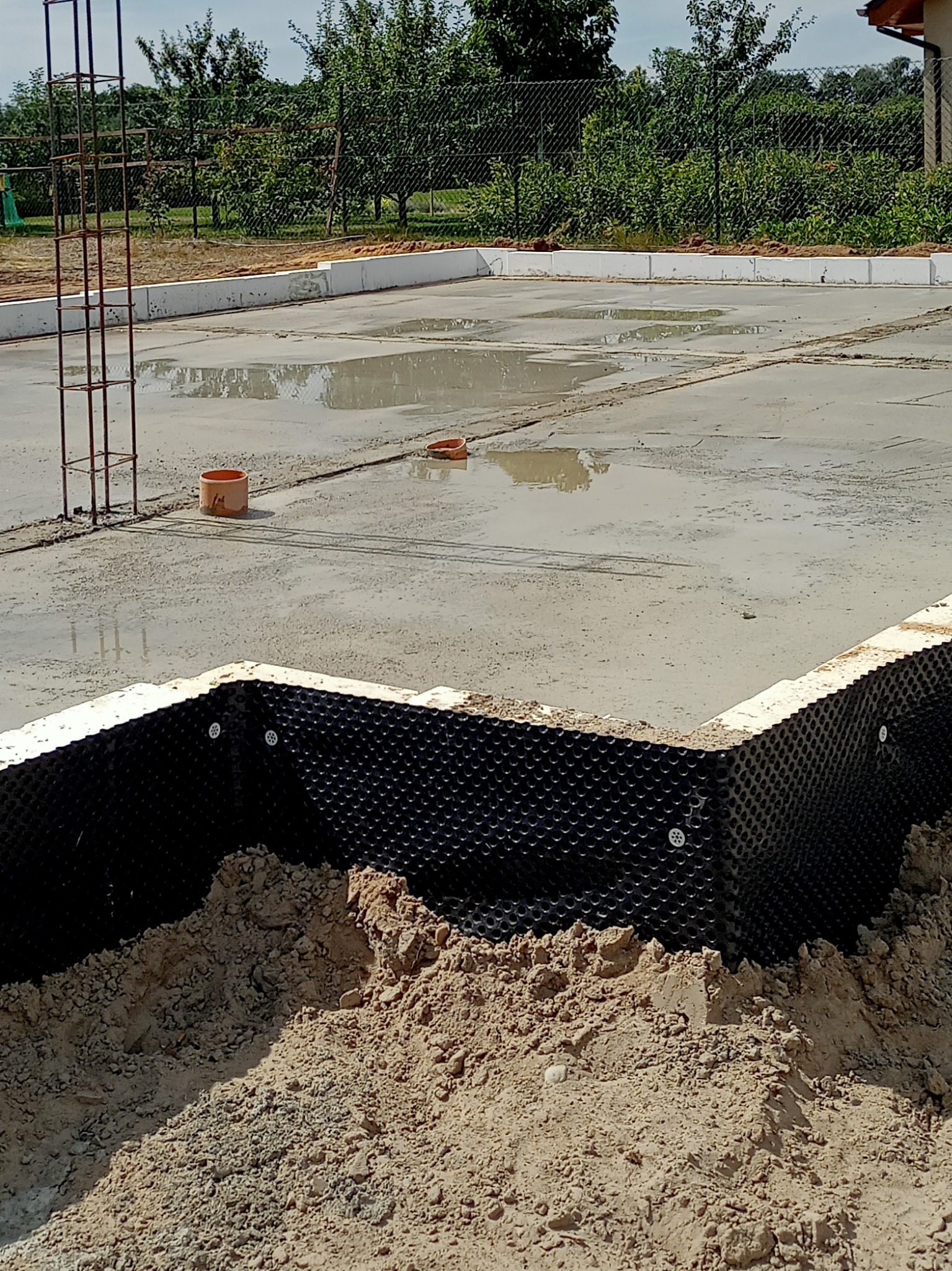 Wylana betonowa płyta fundamentowa z hydroizolacją pionową w postaci folii kubełkowej, widoczne zbrojenie słupów i fragmenty rur kanalizacyjnych, na wierzchu betonowej płyty stoją kałuże wody.