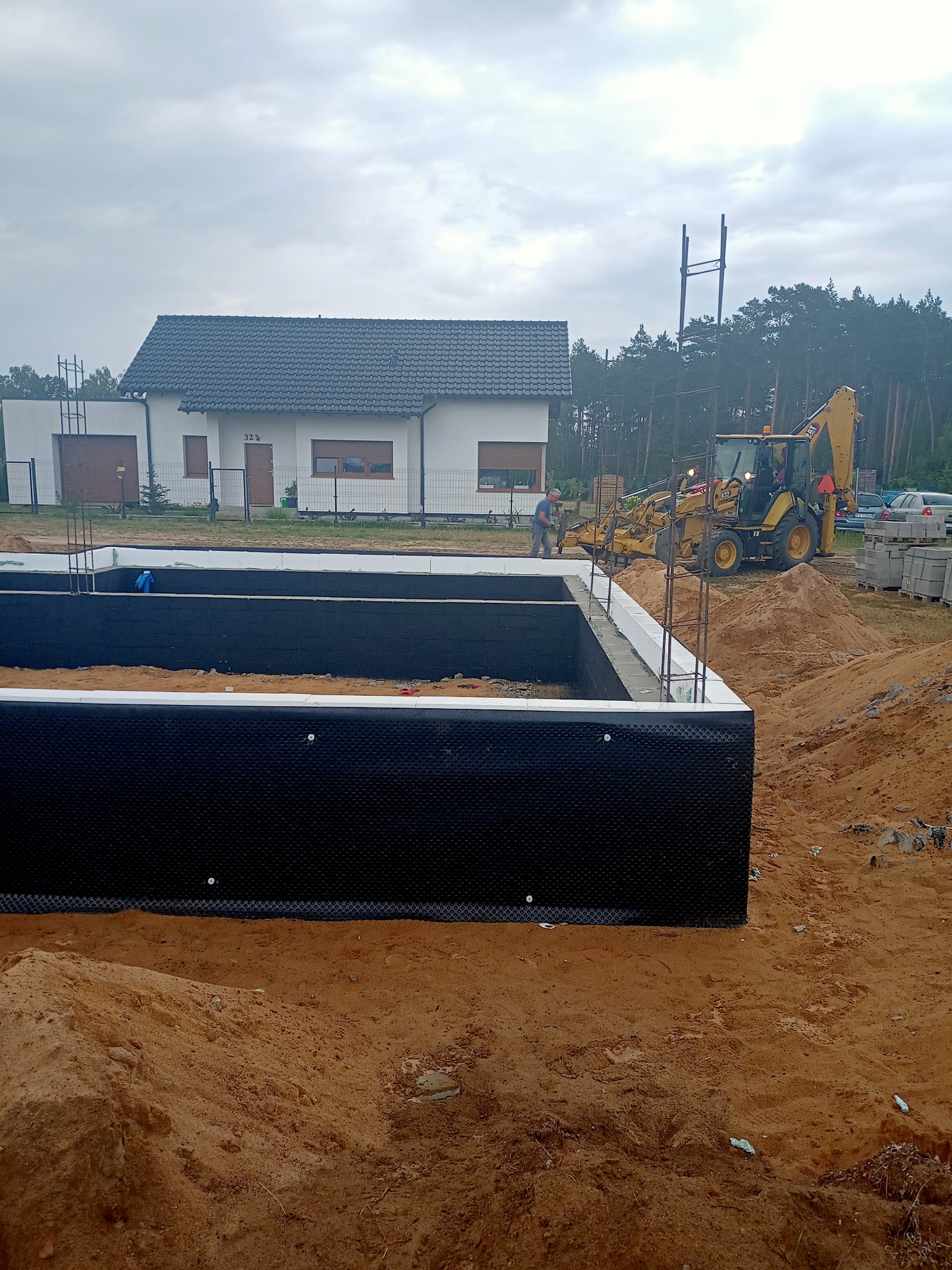 Fundamenty domu jednorodzinnego w trakcie budowy, zabezpieczone czarną folią kubełkową i izolacją termiczną, na tle wykopu z piasku i budynku w tle, widoczna koparka.