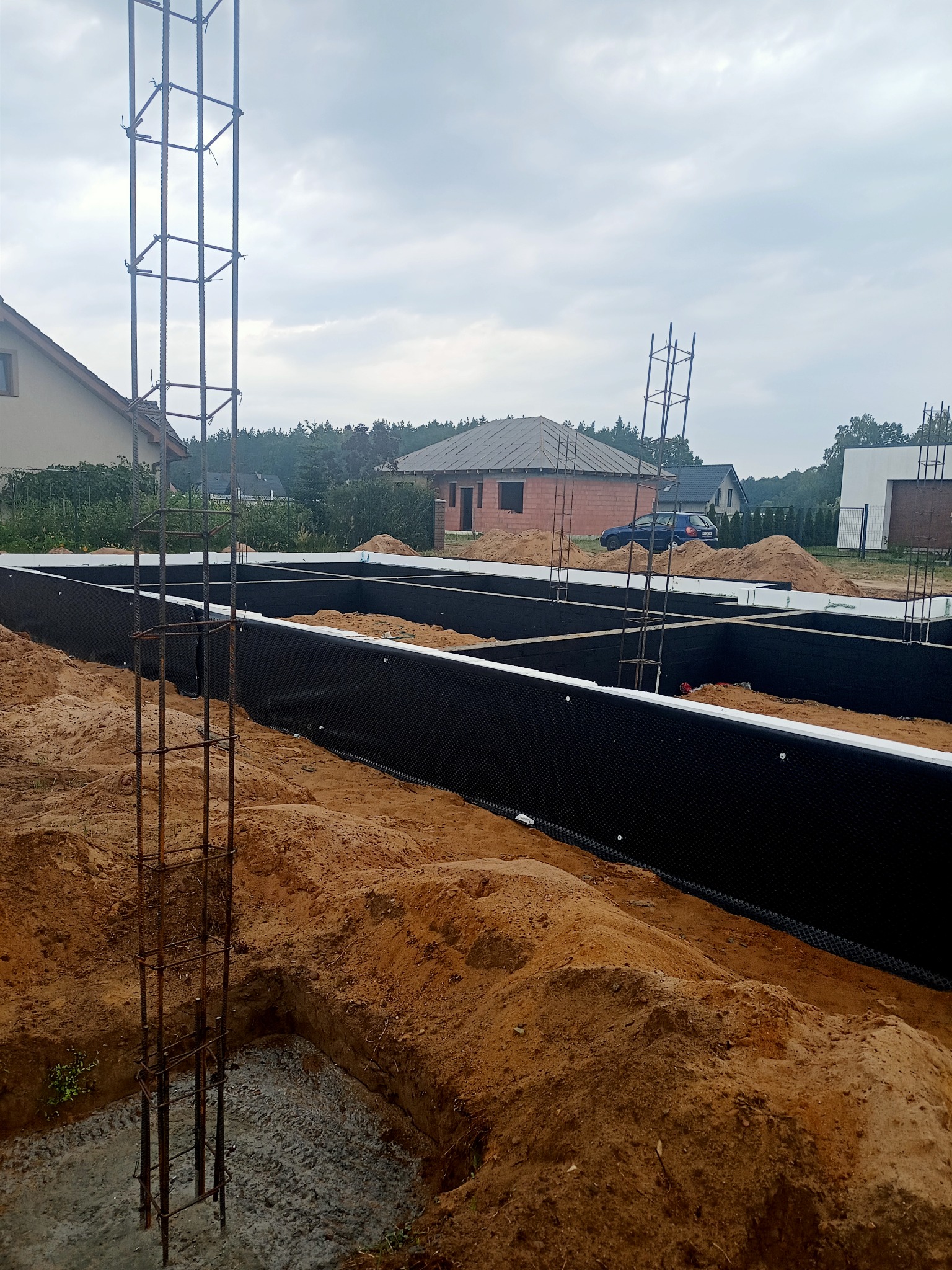 Przygotowanie fundamentów pod budynek: zbrojenie wystające z wykopu, izolacja pionowa z czarnej folii kubełkowej, widoczny piasek i budynki w tle.