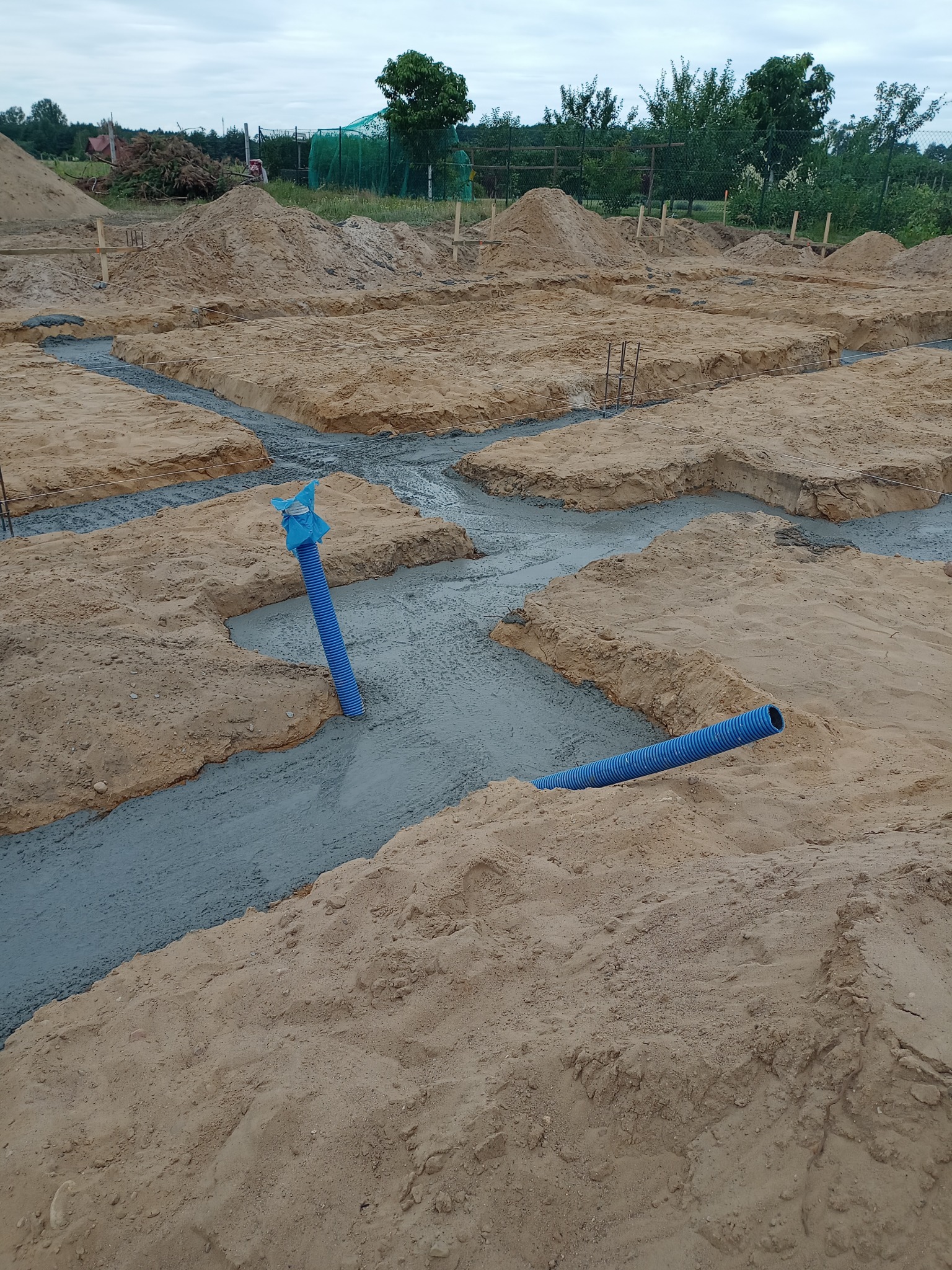 Wylane fundamenty pod budynek z widocznymi niebieskimi rurami instalacyjnymi wystającymi z betonu, otoczone piaskiem i ziemią.