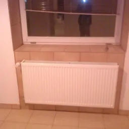 Grzejnik panelowy zamontowany pod oknem z beżowymi płytkami na parapecie i ścianie, widoczne beżowe płytki podłogowe.