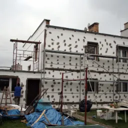 Dwupiętrowy budynek w trakcie ocieplania styropianem, widoczne rusztowania, pracownik przy oknie, porozrzucane narzędzia i materiały budowlane na trawniku, pochmurne niebo.