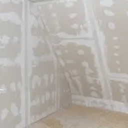 Wnętrze poddasza w trakcie adaptacji: ściany i skosy obłożone płytami gipsowo-kartonowymi, widoczne szpachlowanie łączeń i wkrętów, okno zabezpieczone folią ochronną.