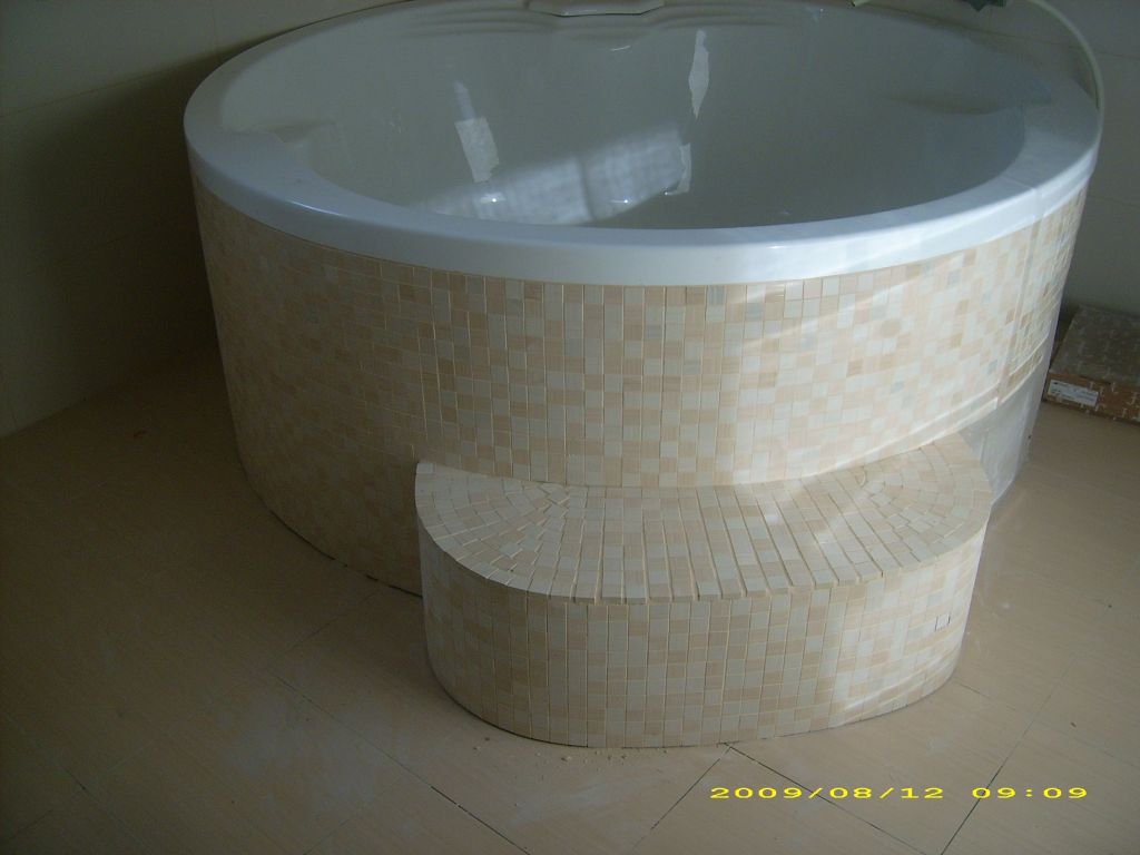 Zaokrąglona wanna z hydromasażem obłożona mozaiką w odcieniach beżu i kremu, z półokrągłym stopniem również wykończonym mozaiką, na jasnej podłodze.