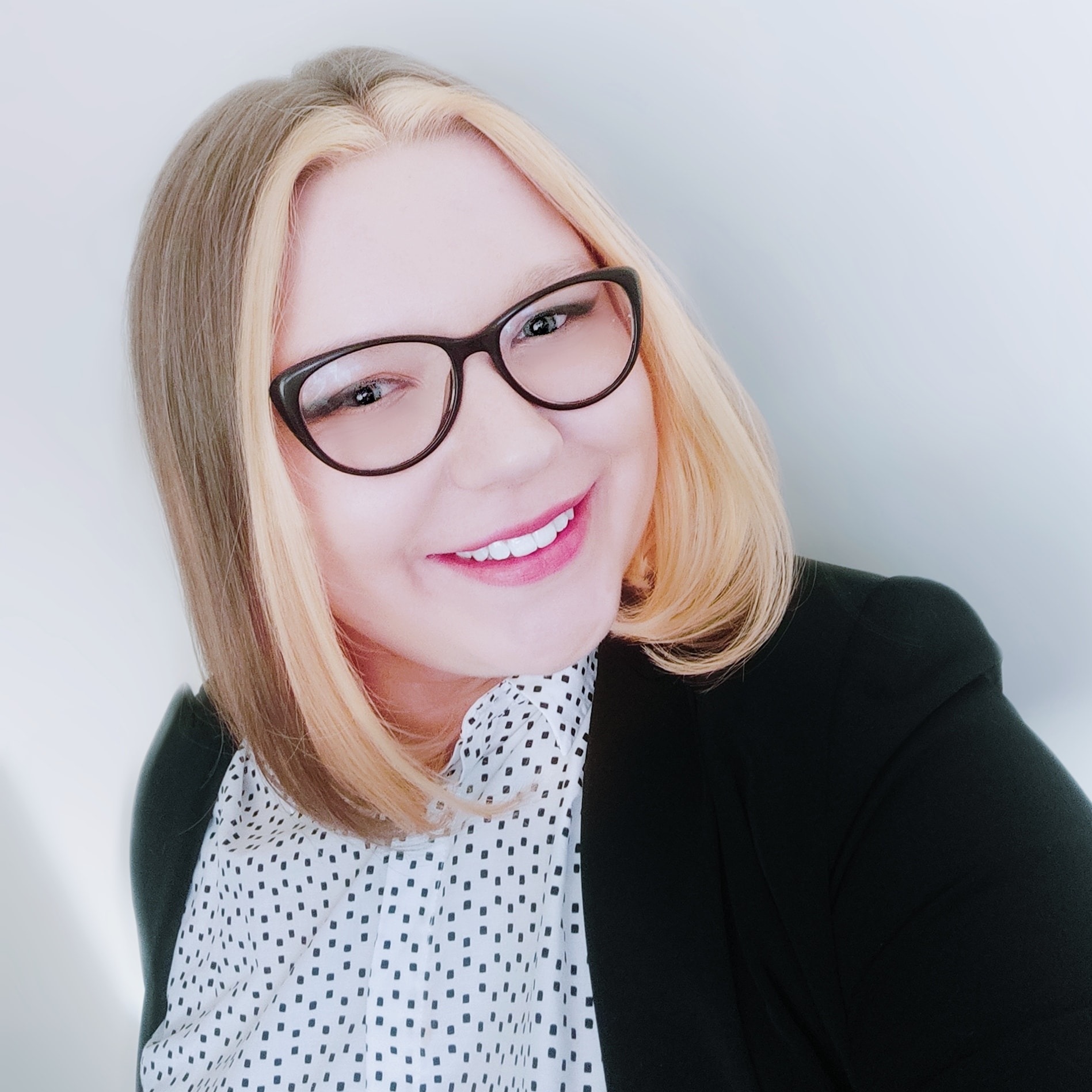Uśmiechnięta kobieta w okularach i marynarce, z blond pasemkami, patrzy w obiektyw.
