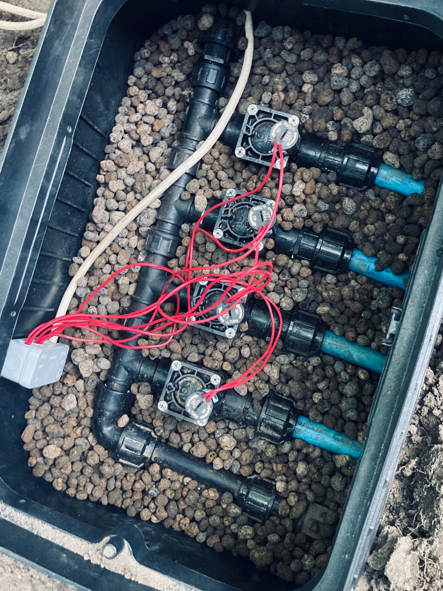 Skrzynka zaworowa z czterema elektrozaworami, przewodami elektrycznymi i rurami doprowadzającymi wodę, wypełniona drobnymi kamykami.