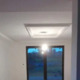 Wnętrze pokoju po odświeżeniu, z nową ramą sufitową i świeżo pomalowanymi ścianami na biało, widok na okno z ciemnymi ramami.