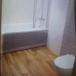 Nowoczesna łazienka z drewnopodobną podłogą, białą wanną z parawanem nawannowym i podwieszaną toaletą.