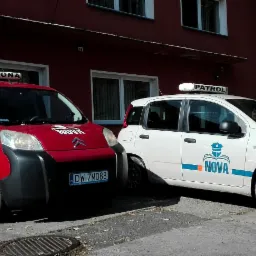 Dwa pojazdy z oznaczeniami agencji ochrony: czerwony Citroen Berlingo z napisem 'Ochrona' i biały Fiat Panda z napisem 'Patrol', zaparkowane przed budynkiem z szyldem agencji.