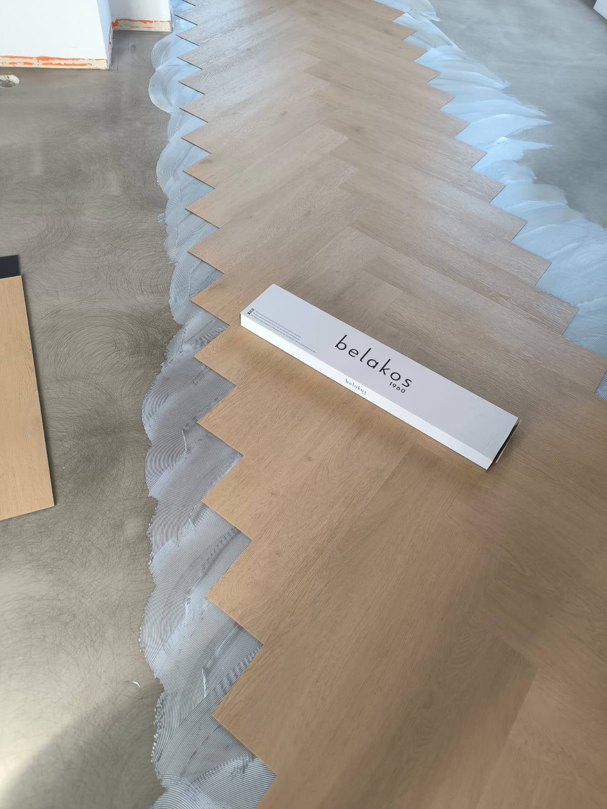 Układanie paneli w jodełkę. Widoczny klej, panele, oraz pudełko z logo Belakos. Jasne, ciepłe odcienie drewna. Etap montażu podłogi.