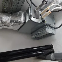 Centrala wentylacyjna z filtrem, podwieszona pod sufitem w pomieszczeniu technicznym, z widocznymi przewodami wentylacyjnymi w izolacji i rurami instalacyjnymi.