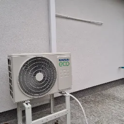 Zewnętrzna jednostka klimatyzacji Kaisai Eco zamontowana na metalowym stelażu przy ścianie budynku z widocznymi elementami instalacji.