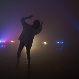 cake2make - Sylwetka osoby w białej koszuli tańczącej w zadymionym pomieszczeniu oświetlonym przez kolorowe reflektory sceniczne.