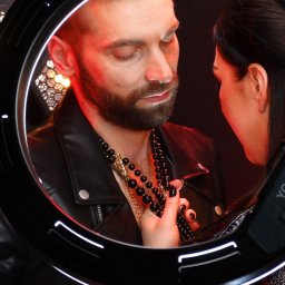 cake2make - Mężczyzna z brodą w skórzanej kurtce i naszyjnikach, widziany przez okrągłą lampę oświetleniową, kobieta dotyka jego naszyjnika, czerwone światło w tle.