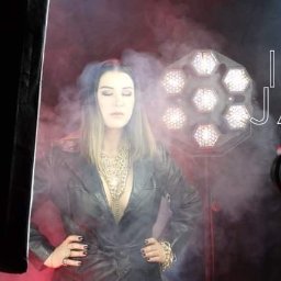 cake2make - Modelka w skórzanym płaszczu i złotej biżuterii pozuje na tle sceny z oświetleniem LED i dymem, widoczny sprzęt operatorski, napis INA JAK