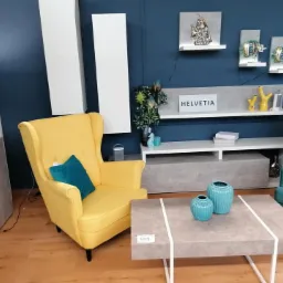 Żółty fotel typu uszak z turkusową poduszką w aranżacji salonu z szarą komodą i stolikiem kawowym. Na ścianie półki z dekoracjami i napis HELVETIA.