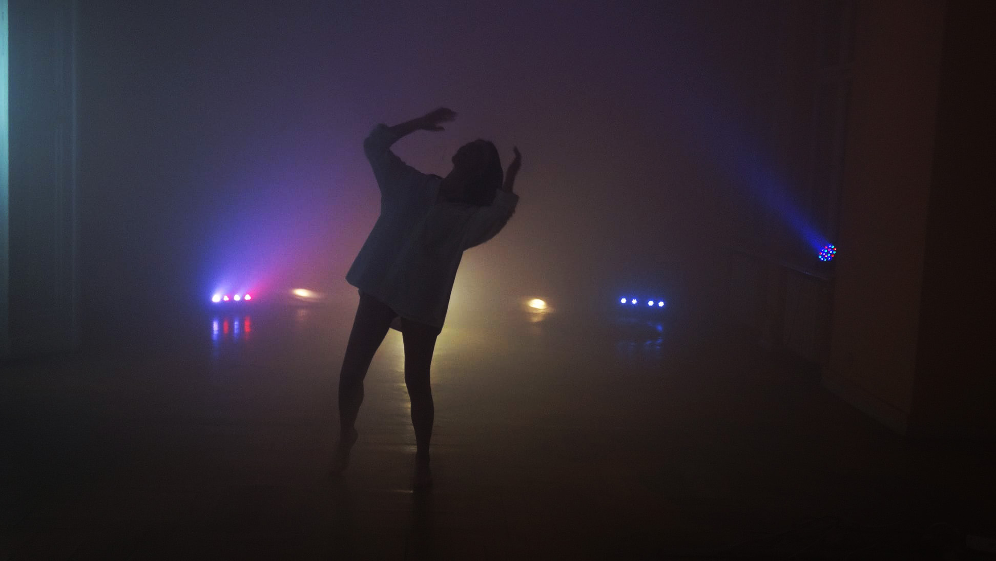 Sylwetka osoby w białej koszuli tańczącej w zadymionym pomieszczeniu oświetlonym przez kolorowe reflektory sceniczne.