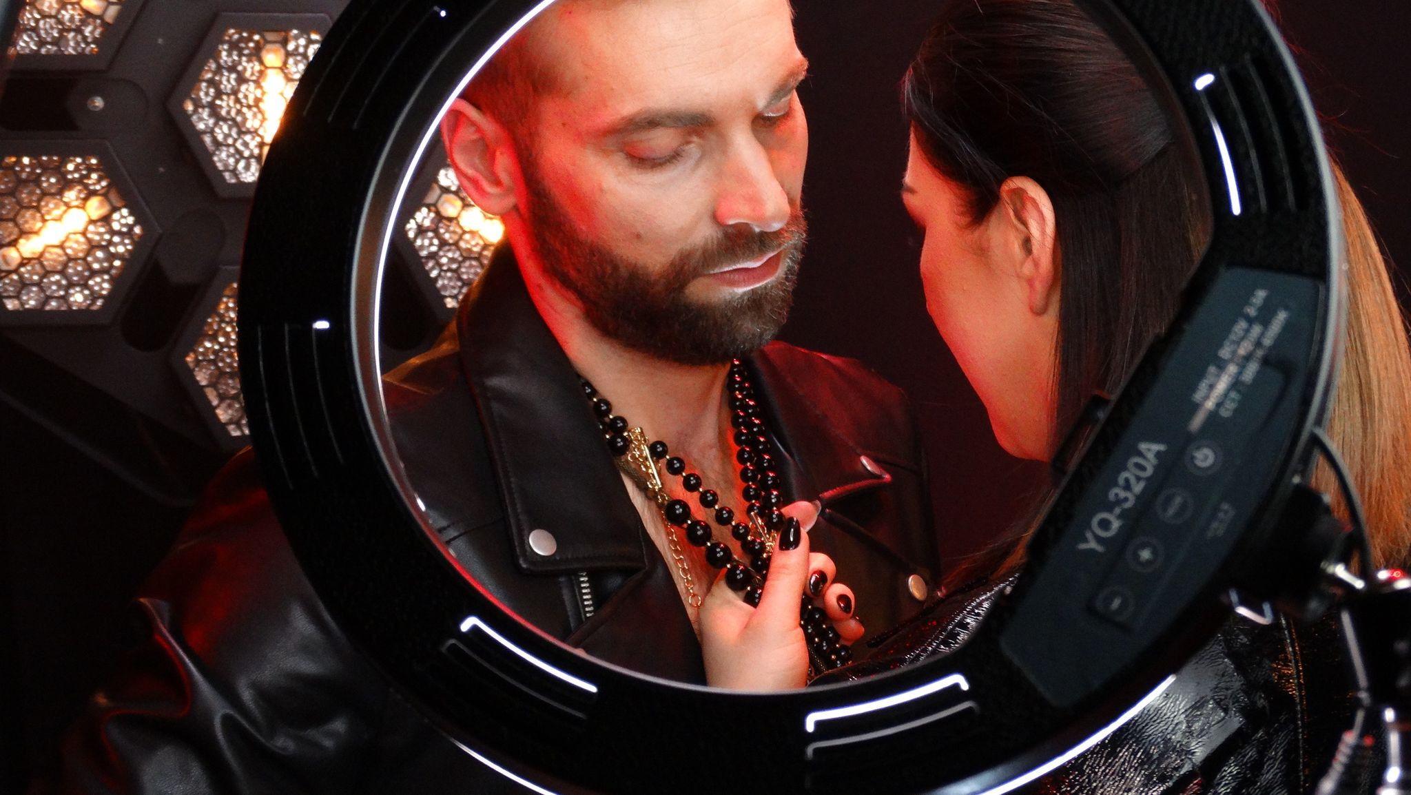 Mężczyzna z brodą w skórzanej kurtce i naszyjnikach, widziany przez okrągłą lampę oświetleniową, kobieta dotyka jego naszyjnika, czerwone światło w tle.