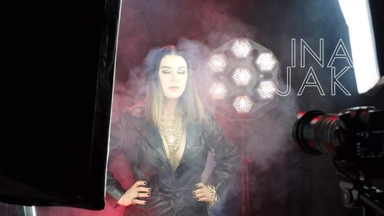 Modelka w skórzanym płaszczu i złotej biżuterii pozuje na tle sceny z oświetleniem LED i dymem, widoczny sprzęt operatorski, napis INA JAK