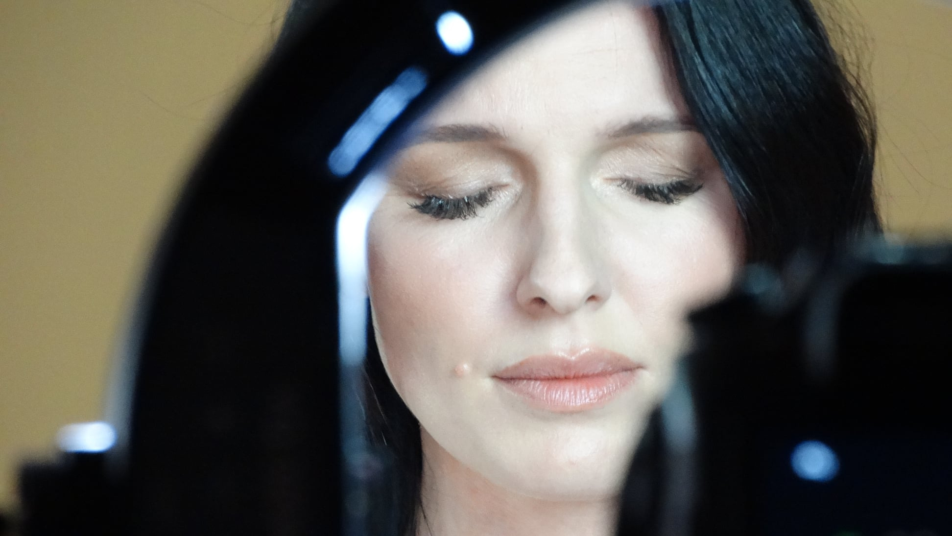 Portret kobiety z makijażem, uchwycony przez obiektyw aparatu z lampą pierścieniową, widoczne sztuczne rzęsy i delikatny błyszczyk na ustach.