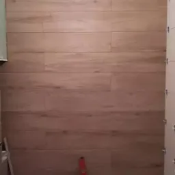 Ściana w trakcie wykańczania płytkami imitującymi drewno, widoczna poziomica i krzyżyki dystansowe.