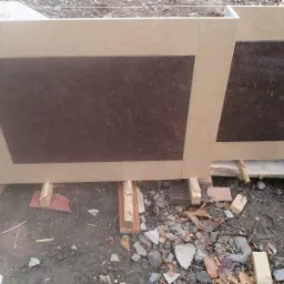 Przygotowane do montażu płyty elewacyjne z brązowym wypełnieniem i beżową ramą, tymczasowo podparte na drewnianych elementach w miejscu budowy.