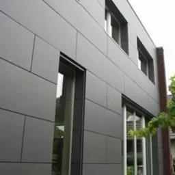 Nowoczesny budynek z szarą elewacją z paneli kompozytowych, widoczne okna z ciemnymi ramami i fragment ceglanego komina.